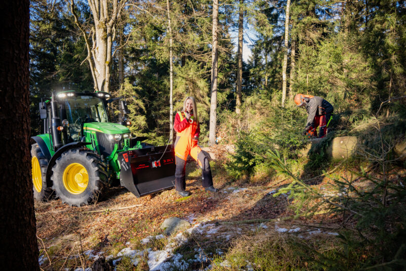 Zwei Personen arbeiten neben einem Traktor in einem Waldgebiet mit Schneeflecken.