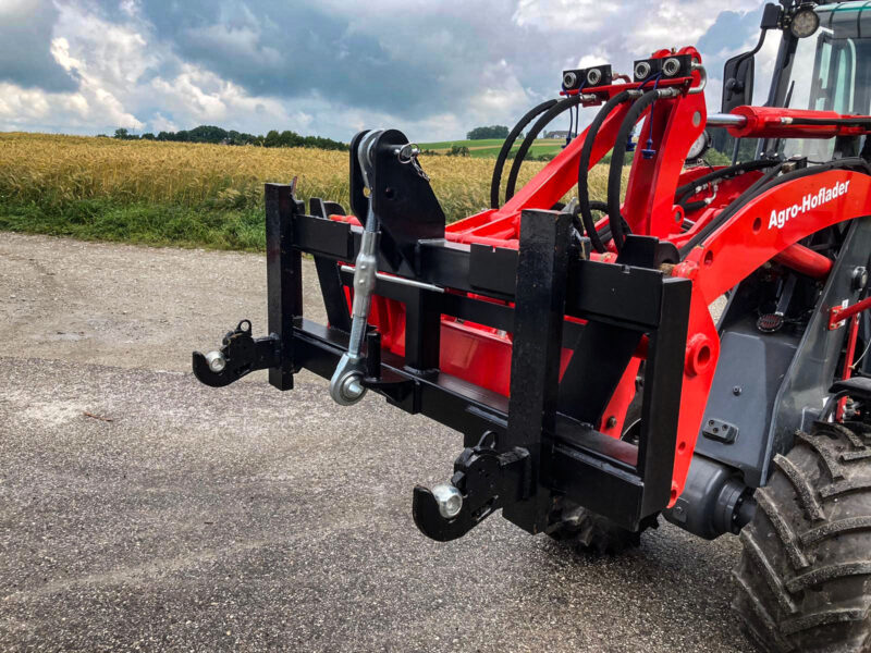 Roter Traktor mit schwarzem Frontanbaugerät, der auf einer Schotterstraße in der Nähe eines Feldes geparkt ist.