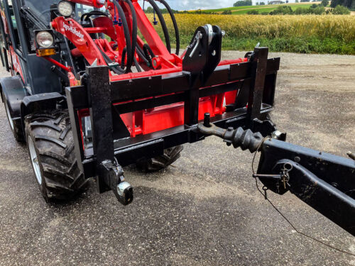 Ein Traktor mit angehängter roter Ausrüstung auf einer asphaltierten Straße in der Nähe von Feldern.