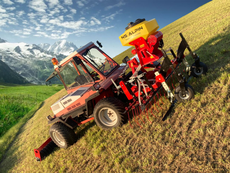 Rotes Fahrzeug auf einem grasbewachsenen Hügel mit Bergen im Hintergrund.